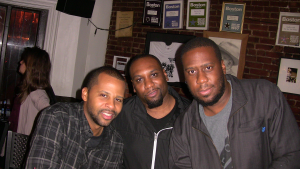 John Roberts, Charles Haynes, and Robert Glasper smiling together inside Wally’s Cafe Jazz Club, with framed awards and photos on the brick wall behind them