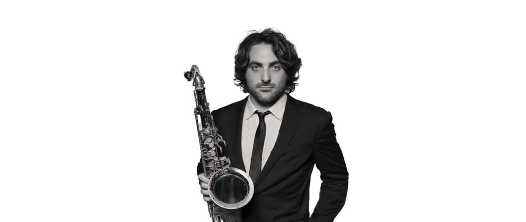 Black and white portrait of saxophonist Noah Preminger holding a tenor saxophone in a suit and tie.