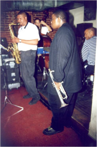 Jeremy Pelt holding a trumpet and Jimmy Greene playing saxophone at Wally’s Cafe Jazz Club with bass and drums in the background.