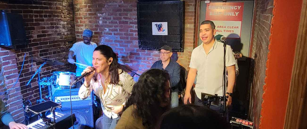 Timba Messengers band performing live at Wally’s Cafe Jazz Club with vocalists, percussionist, and keyboardist in front of an exposed brick wall.